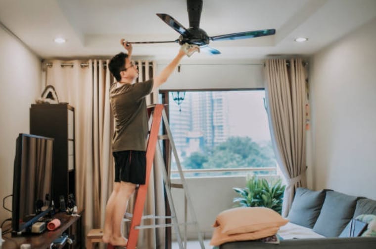 A man cleaning a ceiling fan in a living room