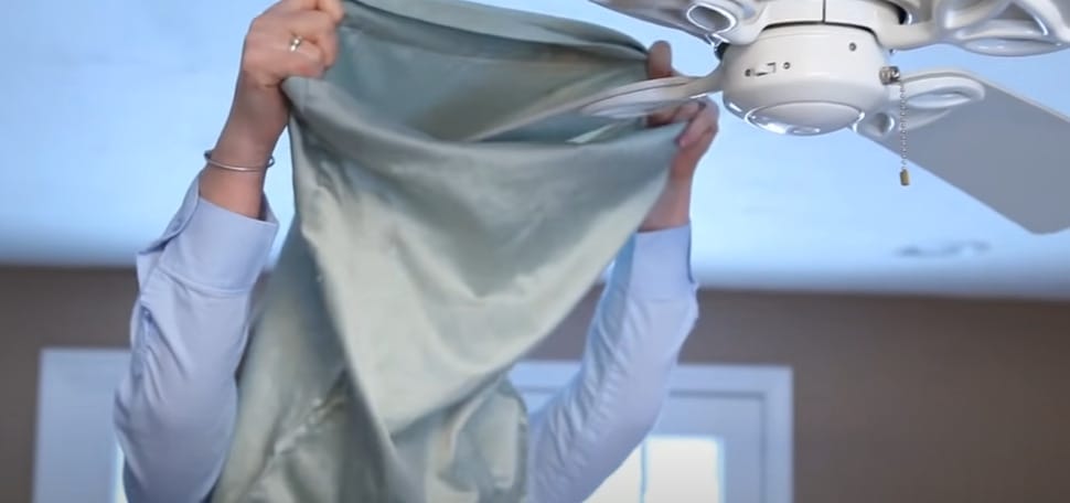 A woman is covering a ceiling fan blade with a pillowcase