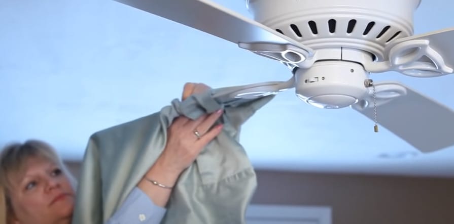 A woman is cleaning a ceiling fan blade with a pillowcase