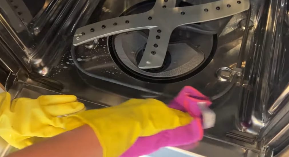 A person in yellow gloves cleaning the inside of a dishwasher using a pink clothe