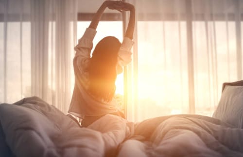 A woman is stretching in bed at sunrise