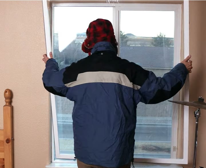 A man in blue, gray black jacket, putting a weather strip on the window