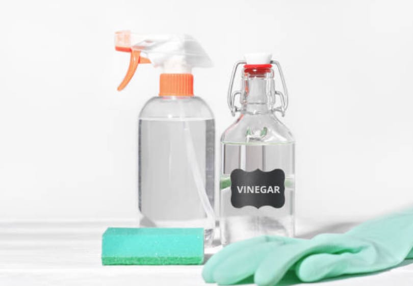 A bottle spray of water, a bottle of vinegar, a sponge and a green gloves on the table