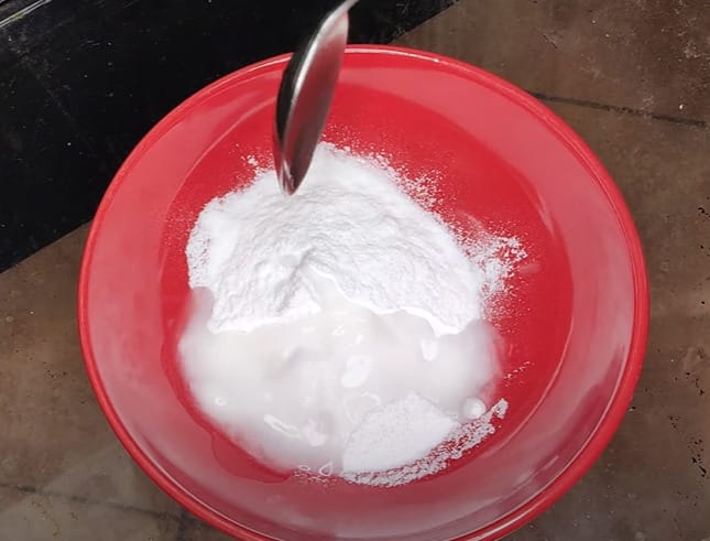 A person putting a spoon of vinegar into a red bowl with baking soda