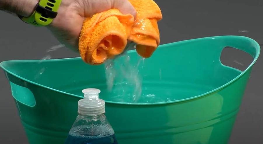 A person squeezing the orange towel from a green bucket with water and liquid detergent besides it