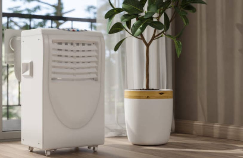 A white portable air conditioner sitting in front of a window besides a pot of indoor plant