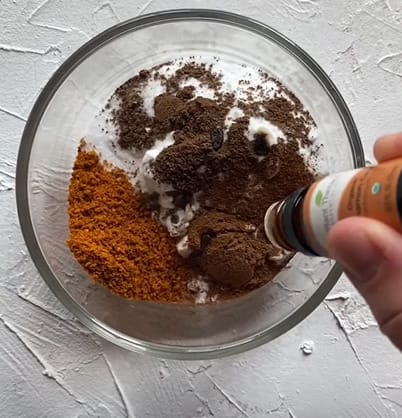 A person pouring cinnamon into a glass bowl of baking soda, essential oil, and rosemary powder
