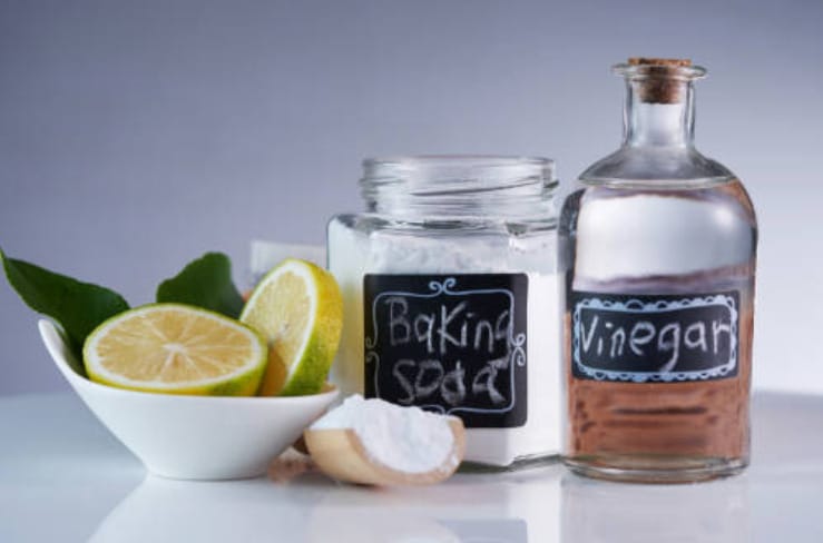 A jar of baking soda, a bottle of vinegar and a bowl of sliced lemon on a table