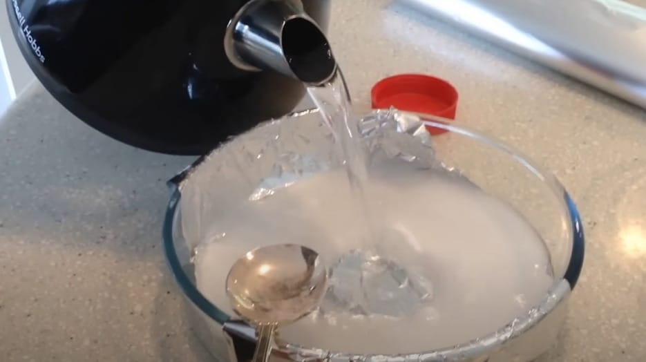 A person putting a hot water into a glass bowl wrapped with aluminum foil and has a baking soda in it