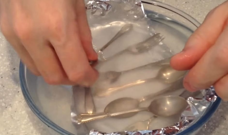 A person placing silverwares on a bowl mixture of baking soda and hot water with aluminum foil