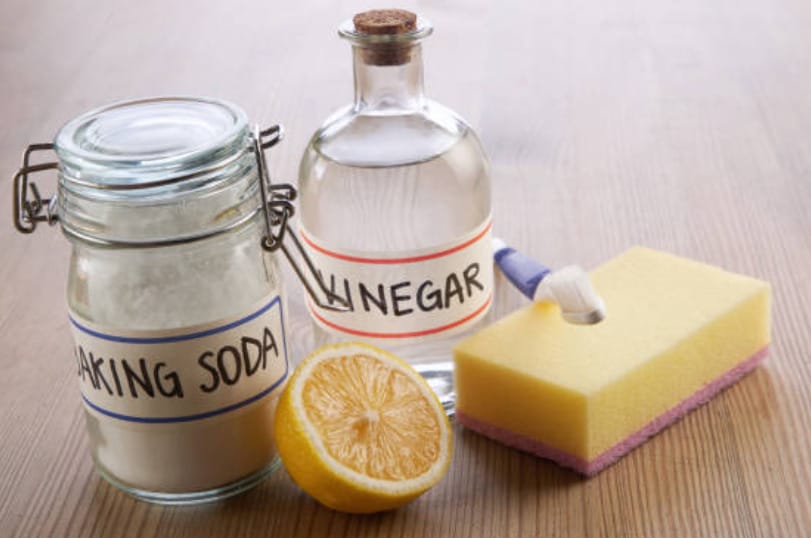A jar of baking soda, a bottle of vinegar, a slice of lemon, a sponge and used toothbrush on a wooden table