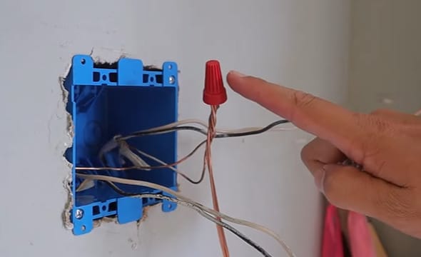 A person is pointing at a red wire nut that is being capped on wire in a blue electrical wall box