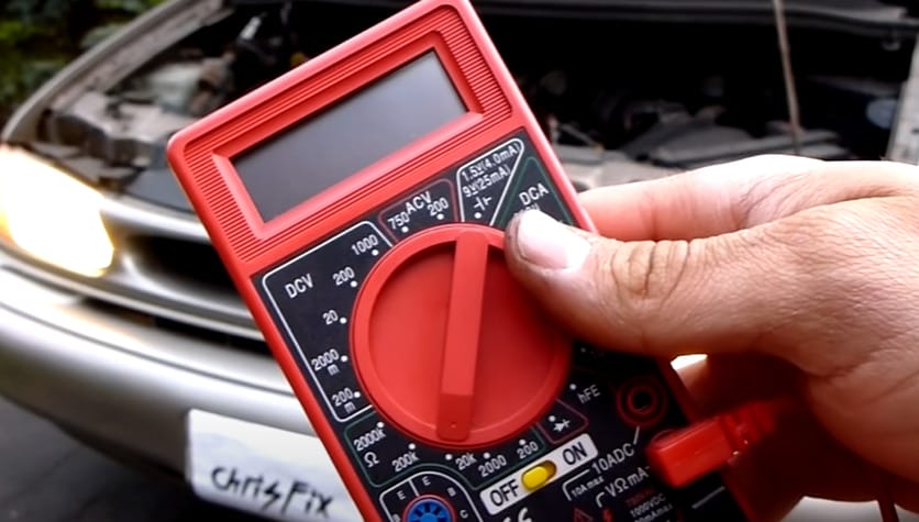 A hand holding a red multimeter and a car with open hood in the background