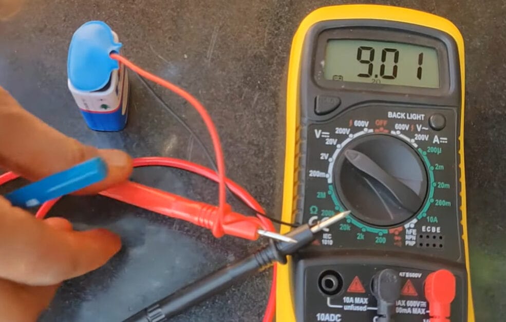 A person holding and pencil and multimeter's probe besides a 12v battery and a multimeter