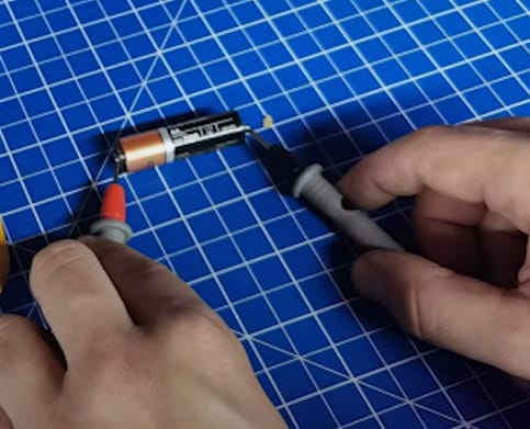 A person is connecting the multimeter's probe to an AA Duracell battery's opposing end