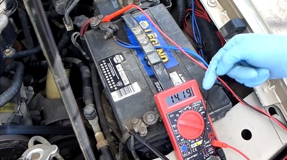 A person is using a multimeter to test a car battery; the multimeter at 14.19 reading