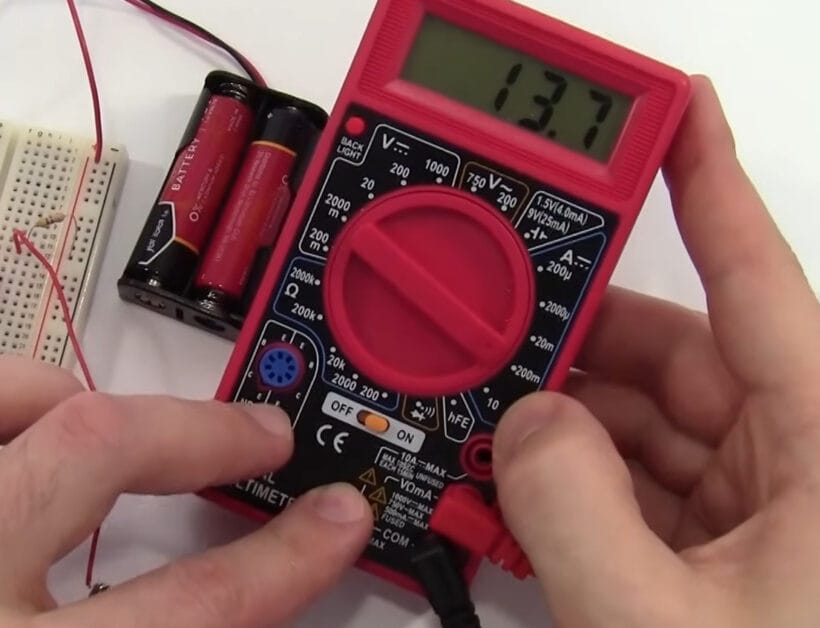 A person is holding a red Cen Tech multimeter and two batteries besides it