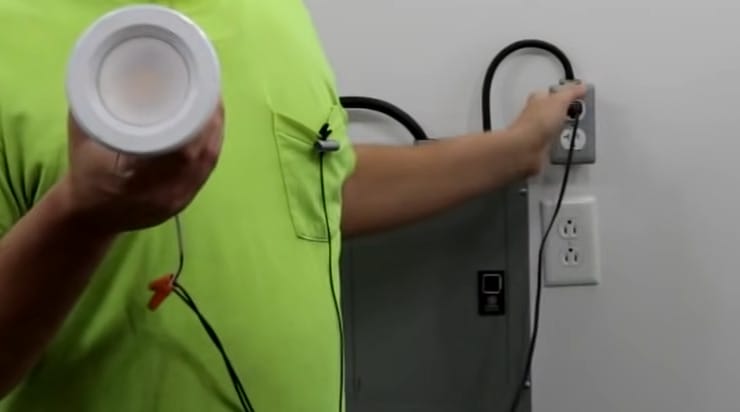 A man in a green shirt is holding a light while performing a test on an electrical outlet with a multimeter