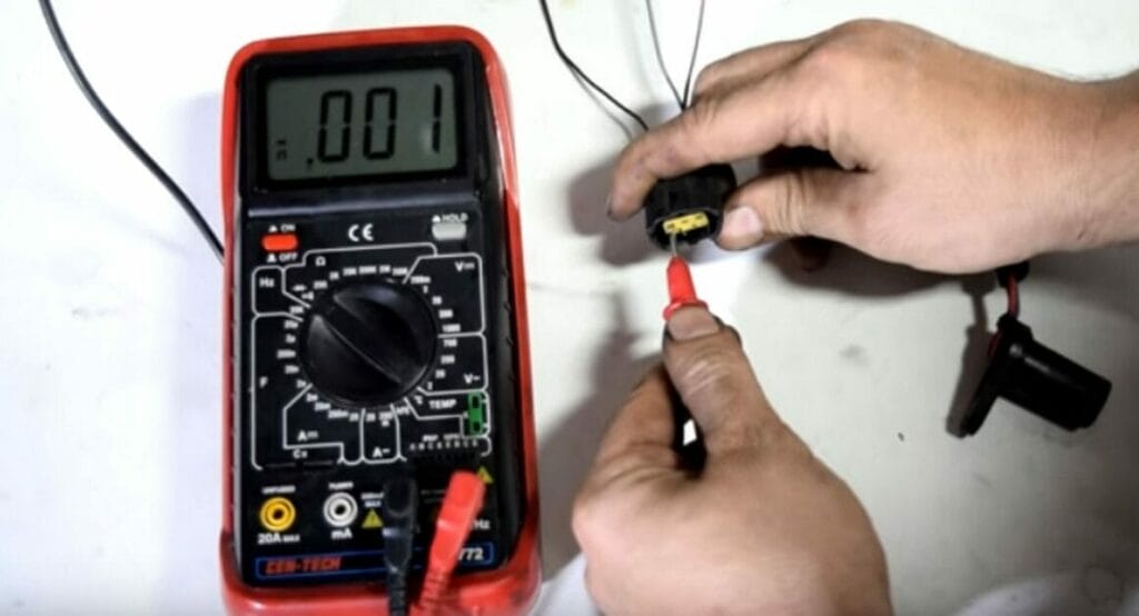 A person is connecting the positive probe to the sensor’s ground wire and the negative one to the battery's negative terminal