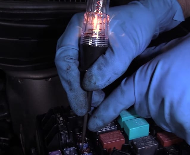 A person in blue gloves is using a test light to test car fuses