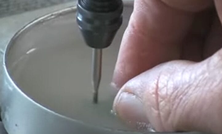 A person using a screwdriver to drill a hole in sea glass underneath a cup of water