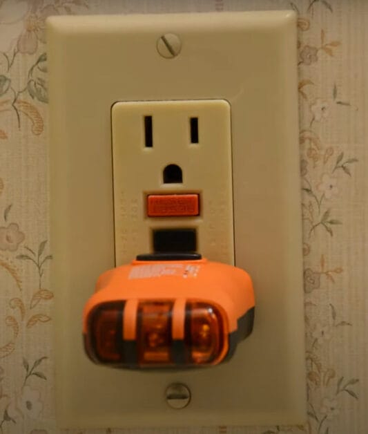 An orange outlet tester is being plugged on the outlet wall