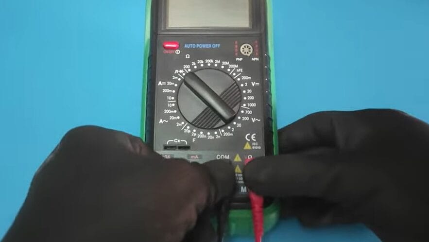 A person demonstrating how to check 240 voltage with a multimeter on a blue background.