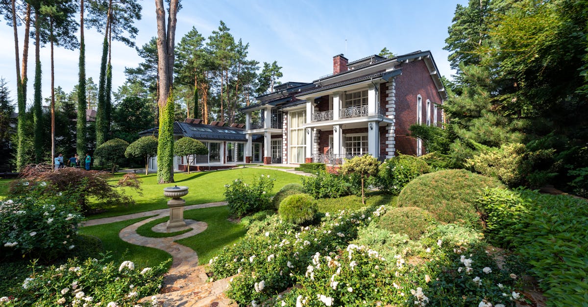 A large, two-story house with pillars and a well-manicured garden featuring a bird bath, surrounded by tall trees and lush greenery.