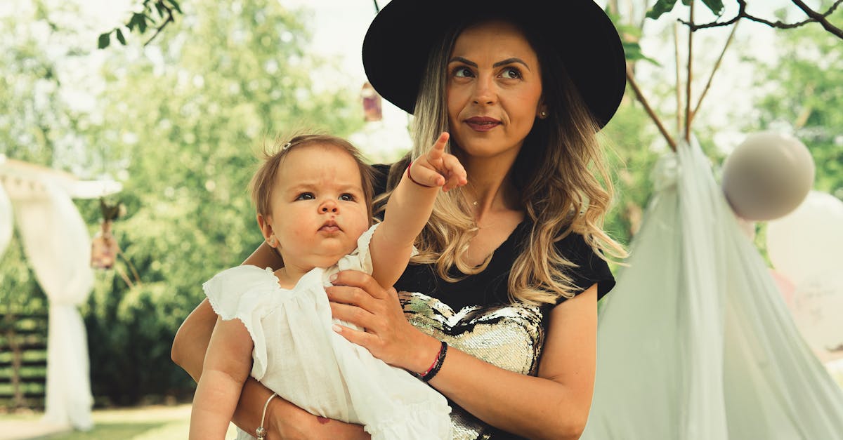 A woman wearing a black hat holds a baby in a white dress, who is pointing forward, in an outdoor setting with greenery in the background.