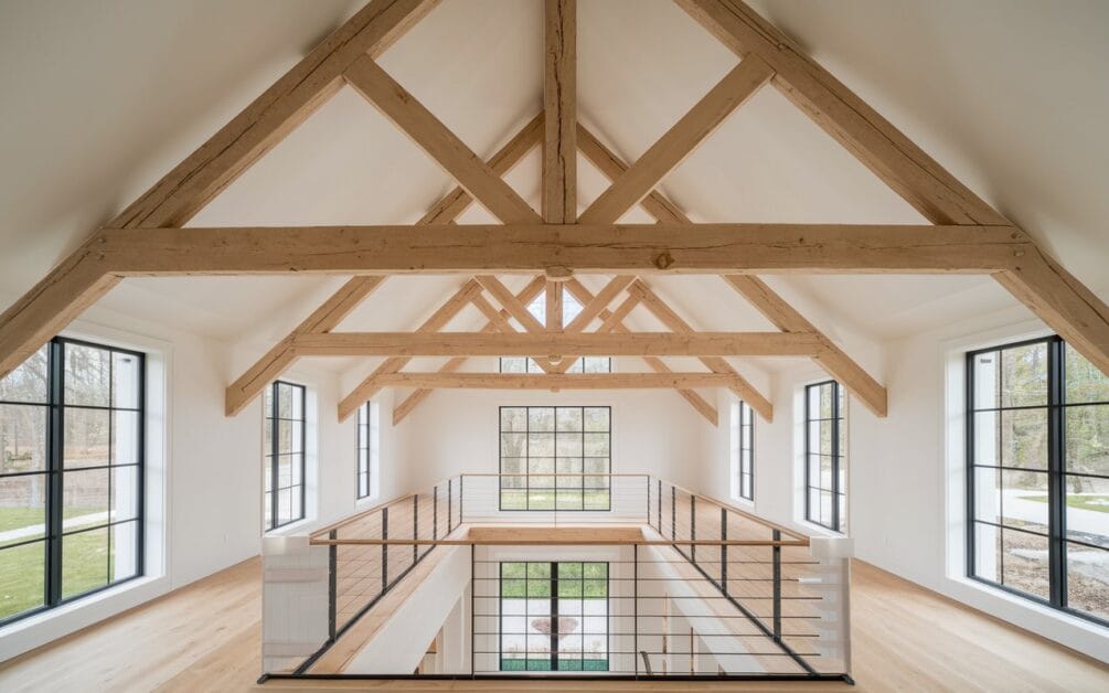 Interior view of a modern room with a high ceiling featuring exposed wooden beams and large windows, providing ample natural light. This Modern Ranch Oasis includes a mezzanine area with a black metal railing, seamlessly blending rustic charm with contemporary elegance.