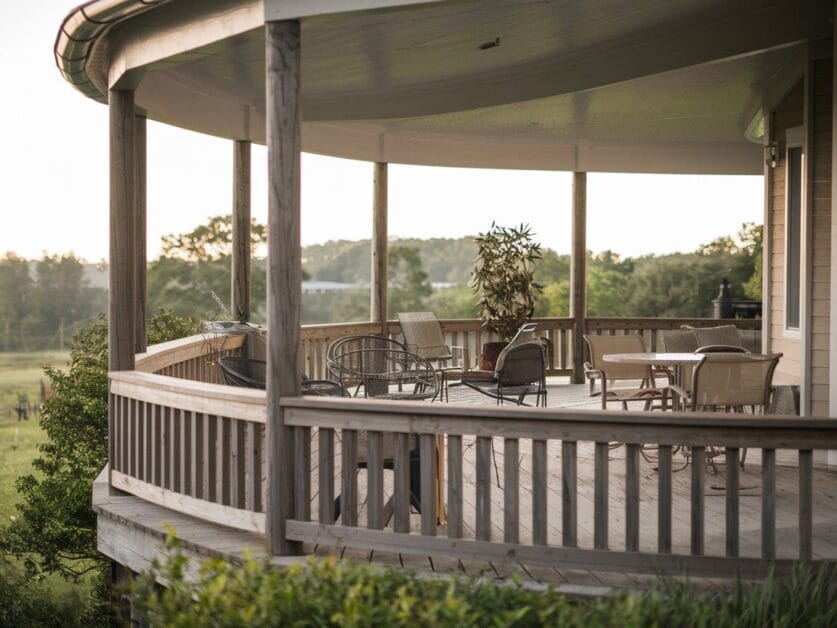 The Whispering Oaks farmhouse boasts a serene wooden deck featuring several empty chairs and tables, with a potted plant in the center. Overlooking a lush green landscape, this partially roofed space invites relaxation and conversation, perfect for enjoying the tranquil surroundings of this 4-bedroom retreat.
