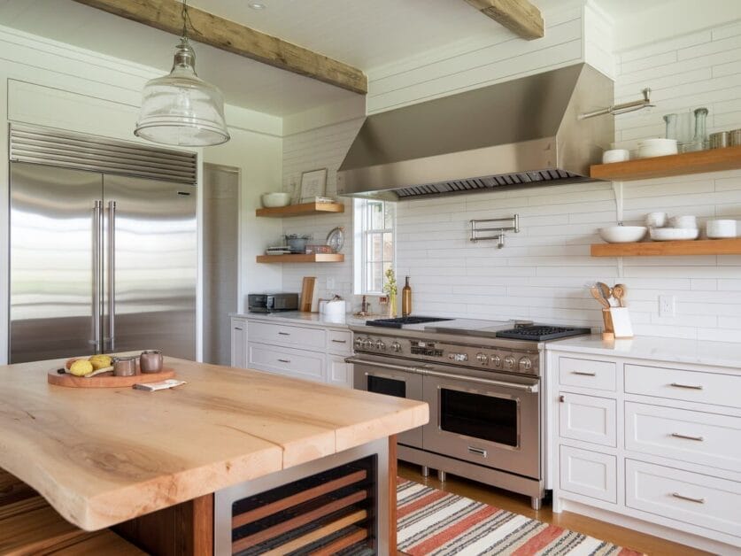 A modern kitchen with stainless steel appliances, a large wooden island, open shelves, and white cabinets. The space features a farmhouse-style design with a wooden beam ceiling, reminiscent of a cozy Red Barn Retreat.