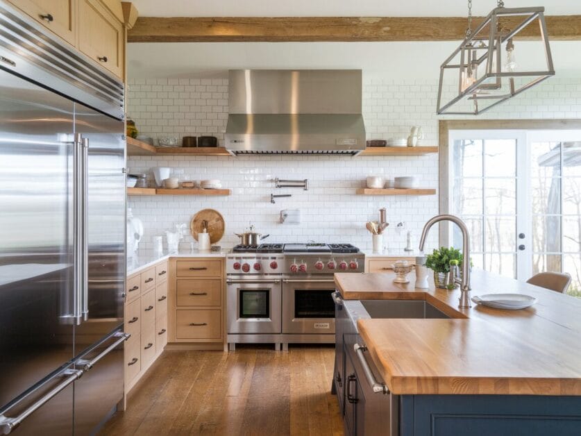 Modern kitchen with stainless steel appliances, wooden cabinets, and open shelves. Center island with wooden countertop and sink embodies farmhouse luxury. Rectangular light fixtures hang from the ceiling, adding a touch of The Midnight Lodge's rustic charm.