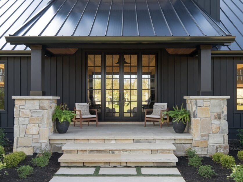 A modern house entrance with a black metal roof, stone steps, and two potted plants. The front boasts two wooden chairs with beige cushions and a large glass-paneled door, exuding a touch of Farmhouse Luxury.