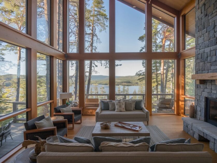 A spacious living room in the Ironclad Cabin, featuring large floor-to-ceiling windows offering a view of a lake and trees, with cozy seating around a stone fireplace. This 4-bedroom retreat is perfect for nature lovers.