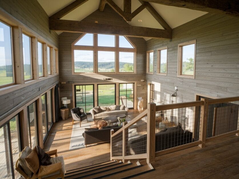 A spacious, open-plan living room with large windows, wooden accents, a high ceiling, and modern furnishings. Set in the dreamy Whispering Oaks, this 4-bedroom home offers panoramic views of the lush green landscape outside.