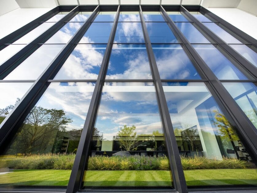 The Ironclad Cabin's large glass windows reflect a blue sky with clouds and surrounding greenery, offering stunning views from the 4-bedroom retreat. A grassy lawn and shrubs are visible in the foreground.