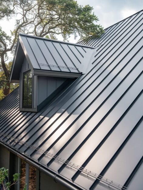 A close-up view of a modern farmhouse with a gray metal roof and a dormer window. Trees are visible in the background, enhancing the retreat-like atmosphere.