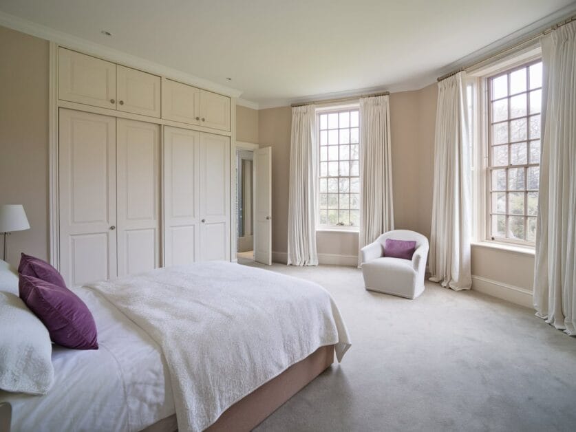 A spacious bedroom with beige walls, large windows with curtains, a bed with white bedding and purple cushions, built-in closets, and a white armchair creates a cozy corner in this 3,500 Sq. Ft. modern farmhouse.