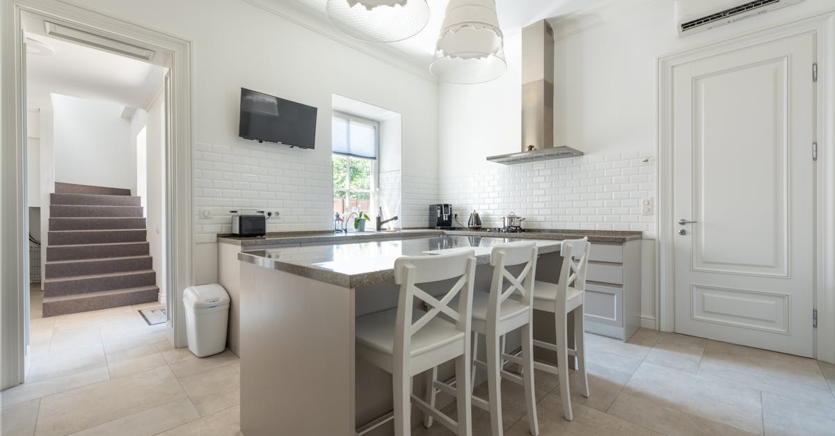 Modern kitchen with white cabinets, a central island with three chairs, stainless steel appliances, and a wall-mounted TV, next to a staircase and door.