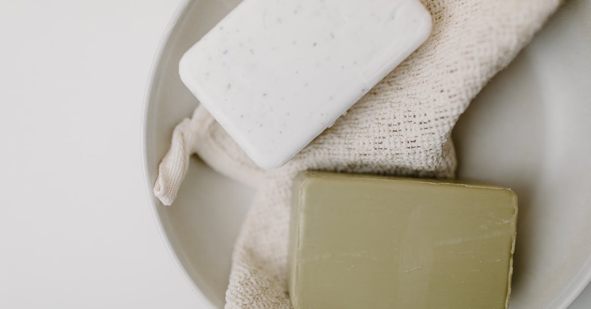Two rectangular bars of soap, one white and one green, rest on a beige textured cloth inside a round white bowl.