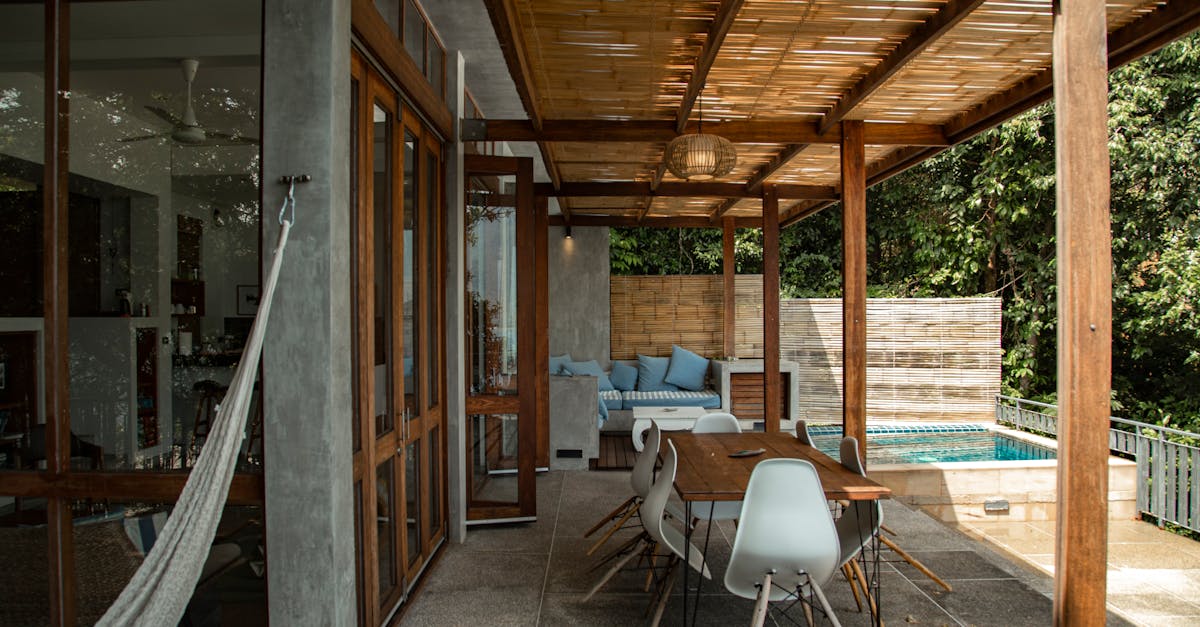 Modern outdoor patio with a wooden pergola, dining table with white chairs, hammock, lounge area with blue cushions, and a small pool surrounded by greenery.