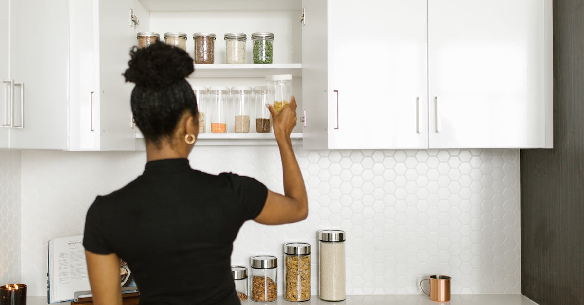 A woman stands in a kitchen reaching for a glass jar on a shelf, with other jars of dry goods on the counter and in the cupboard.
