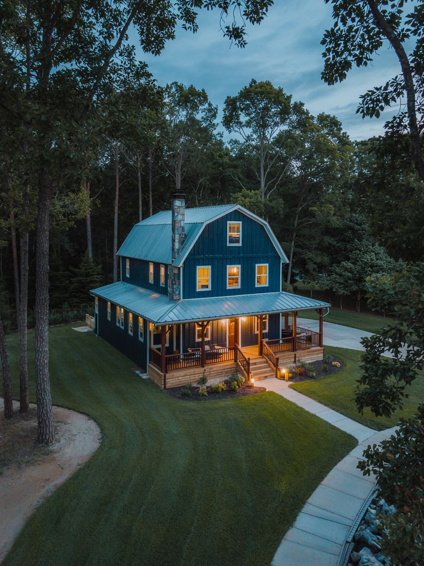 A two-story blue house with a metal roof and wraparound porch is lit up at dusk, surrounded by well-kept lawn, trees, and a curved driveway.