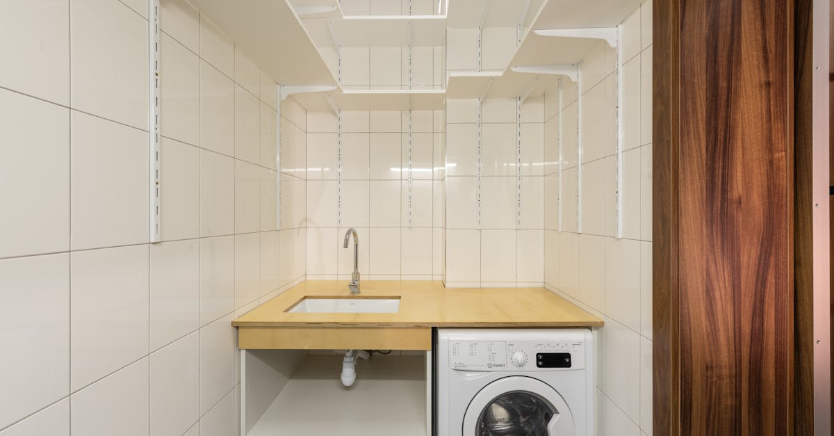 Laundry room with white tiled walls, a wooden countertop with a sink, and a front-loading washing machine underneath. Open shelving brackets are mounted on the walls.