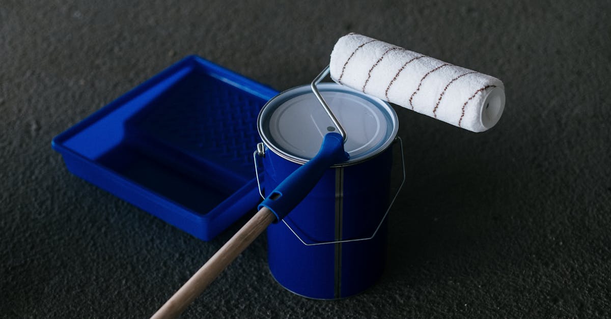 A blue paint tray, a closed blue paint can, and a paint roller with a long handle resting on top, set on a dark surface.