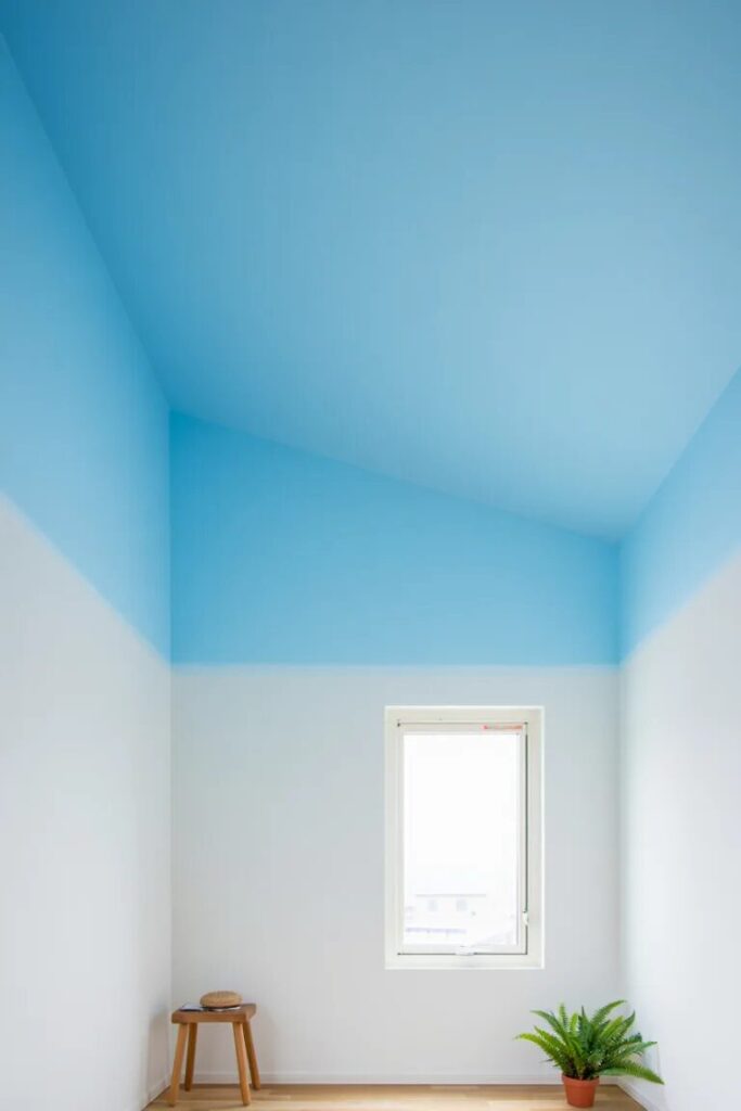 A small, minimal room with white walls, a blue ceiling, a single window, a wooden stool, and a potted plant on the floor.