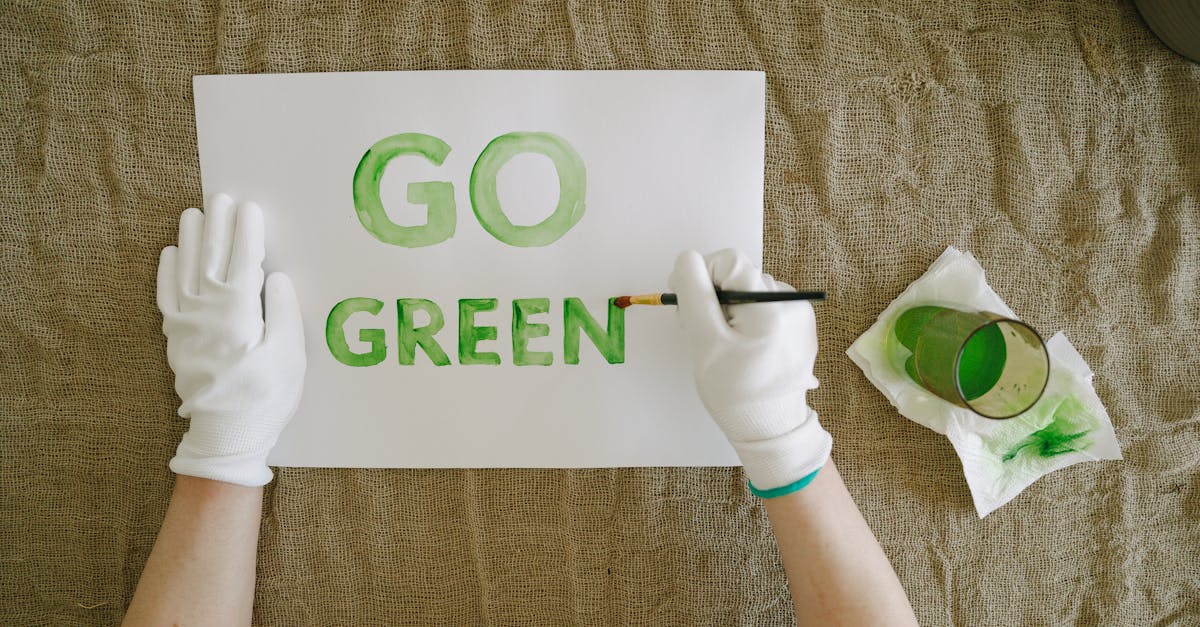A person wearing white gloves paints the words "GO GREEN" in green watercolor on a white sheet of paper, with a paintbrush and a green cup nearby.