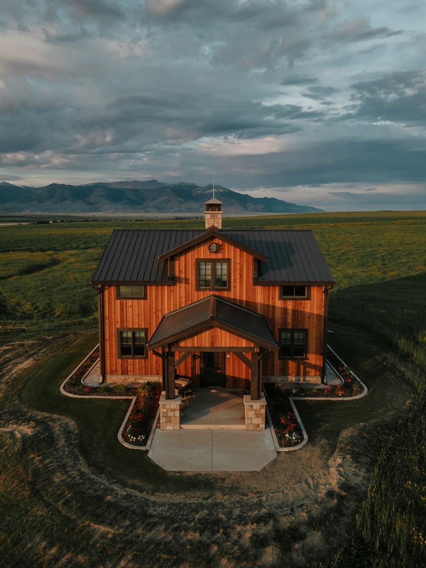 A two-story wooden house with a metal roof sits in a rural field with mountains in the background under a cloudy sky.