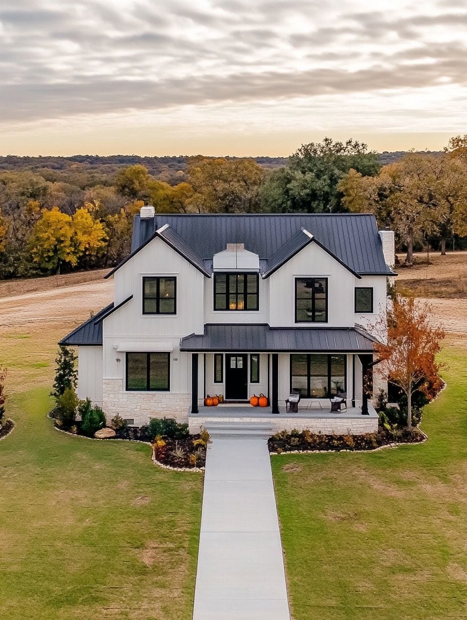 Two-story modern farmhouse with black roof, white siding, large front porch, pumpkins by the entrance, and a concrete walkway, surrounded by grass and autumn trees.